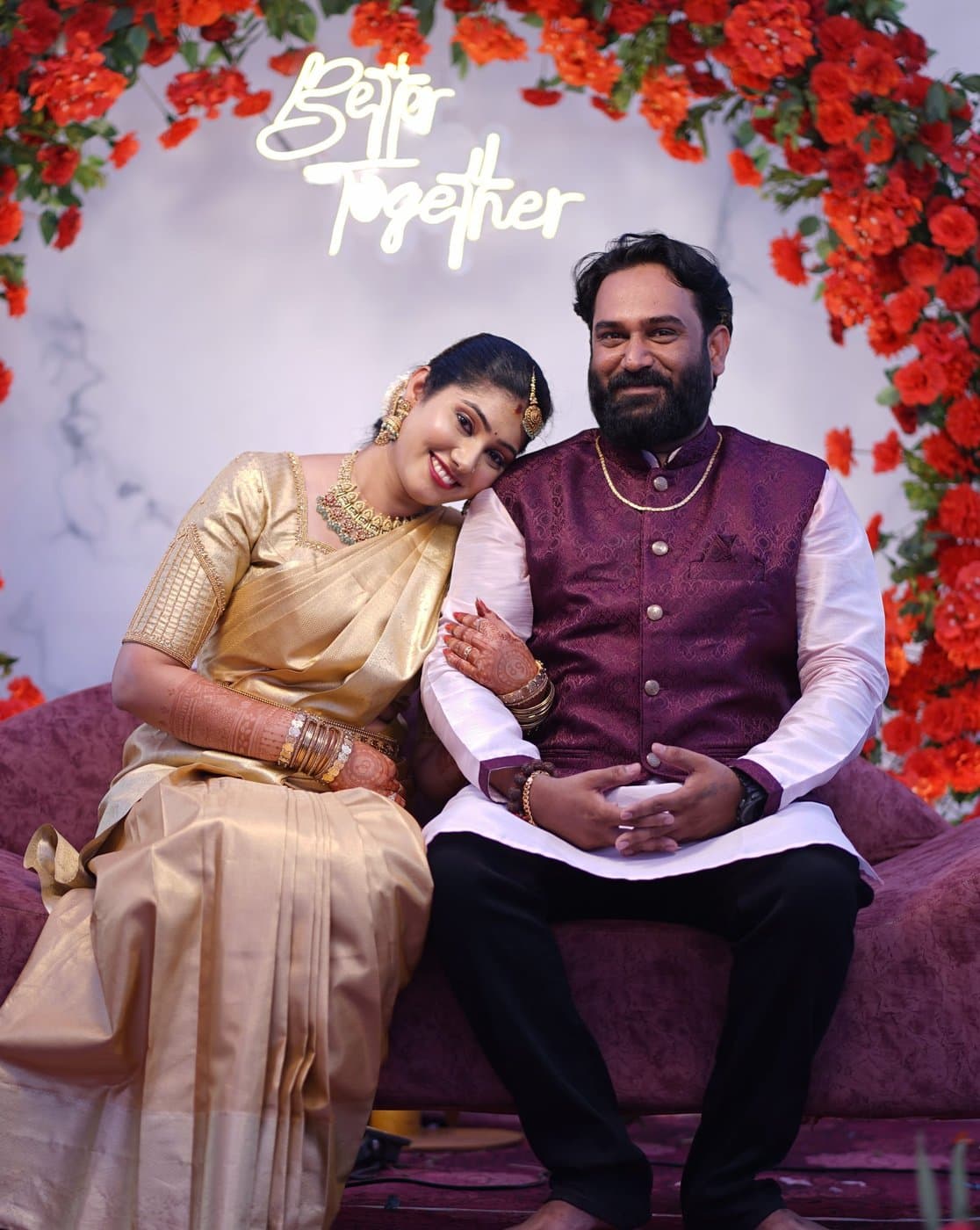 Shravya resting her head on Pramod's shoulder as they sit together on a burgundy sofa under an arch of orange roses